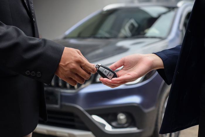 A close-up of a person handing car keys to another individual in front of a vehicle, symbolizing a car purchase or transfer.