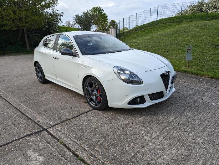 A sleek white Alfa Romeo hatchback parked on a concrete surface, showcasing its stylish design and black alloy wheels against a green background.